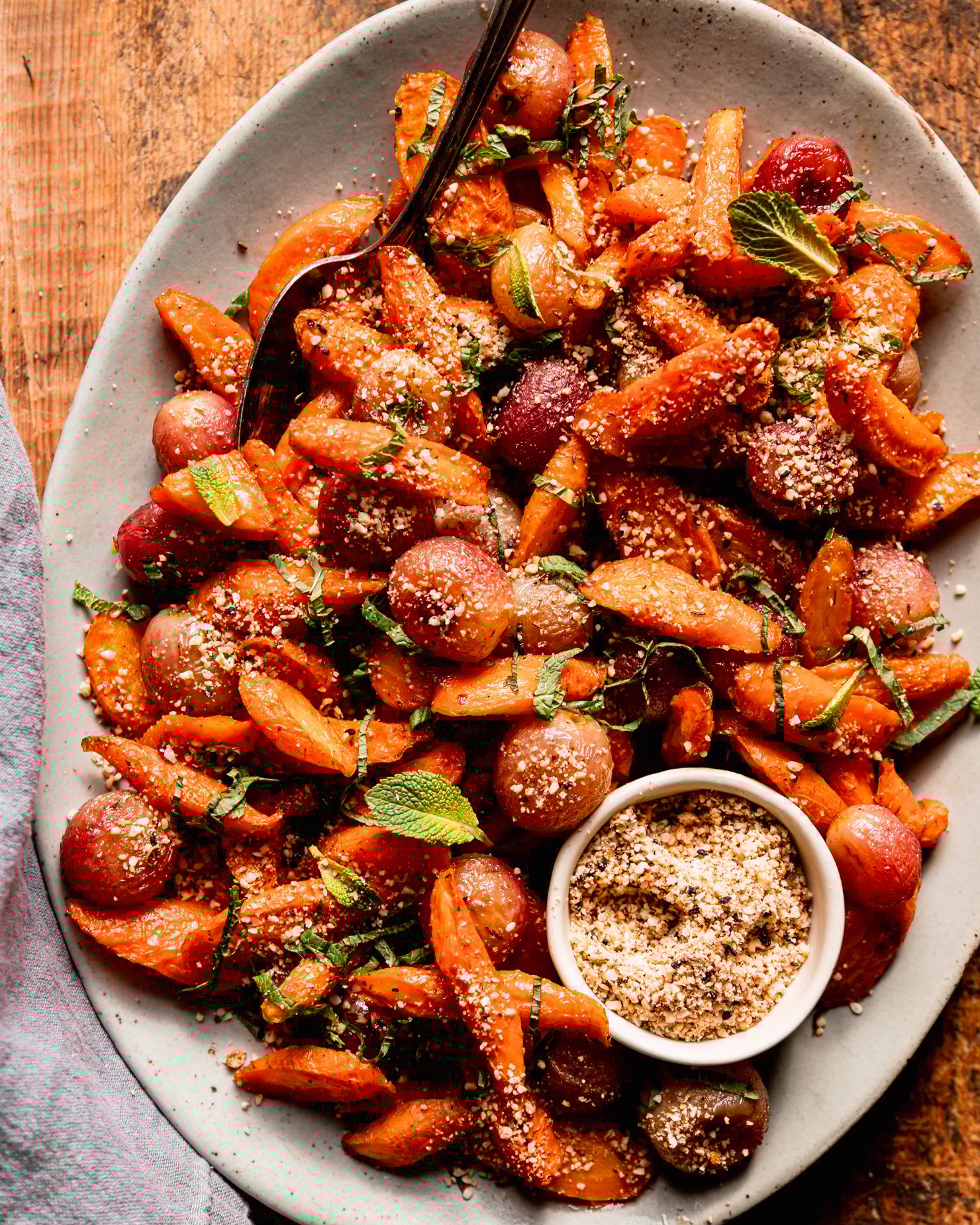 An overhead shot shows a platter of oven roasted carrot side dish featuring roasted grapes, sprinkles of hazelnut-based dukkah, and finely sliced mint.