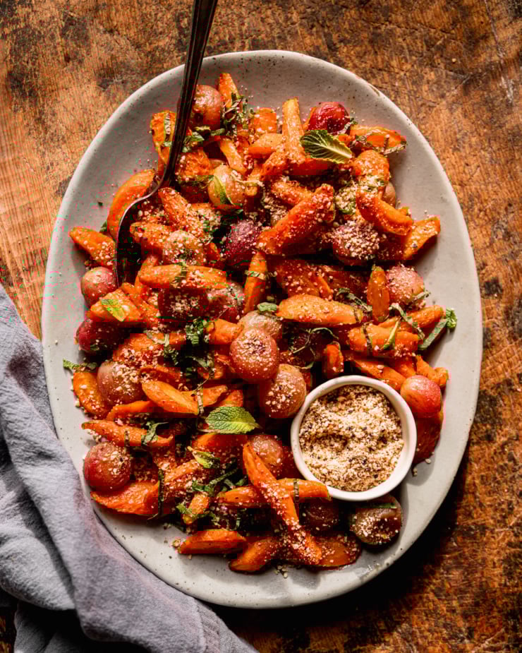 An overhead shot shows a platter of oven roasted carrot side dish featuring roasted grapes, sprinkles of hazelnut-based dukkah, and finely sliced mint. There is a small bowl of dukkah on the platter as well.