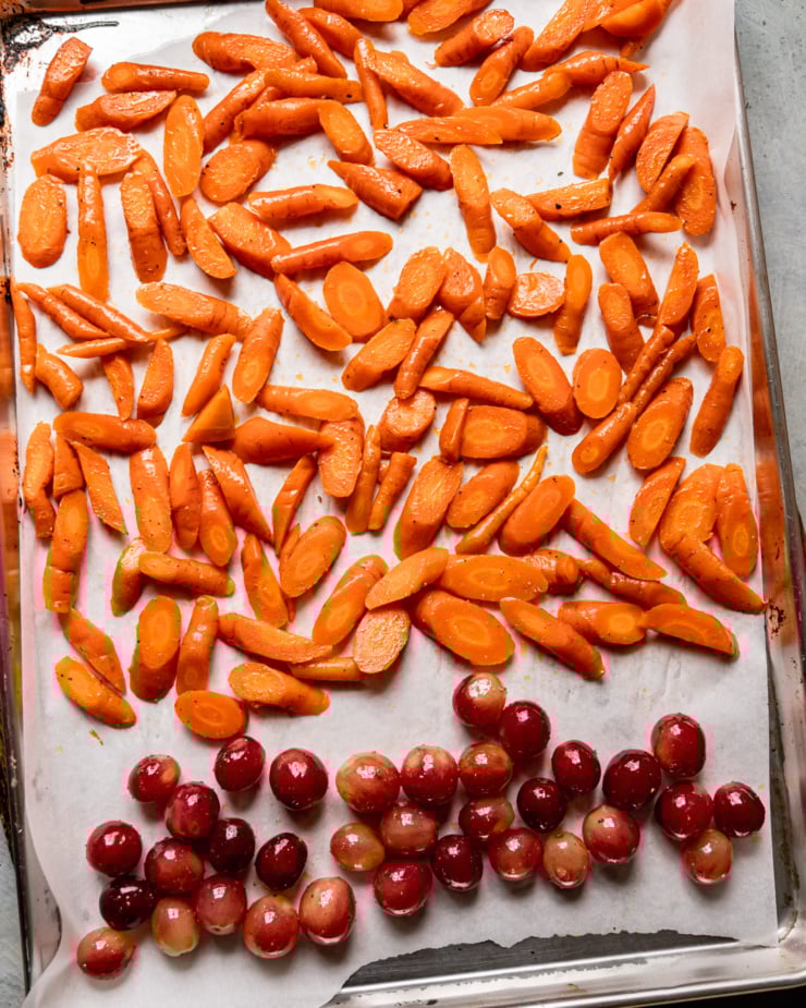 An overhead shot shows chopped carrots and whole red grapes on a baking sheet, coated in olive oil, salt, and pepper.