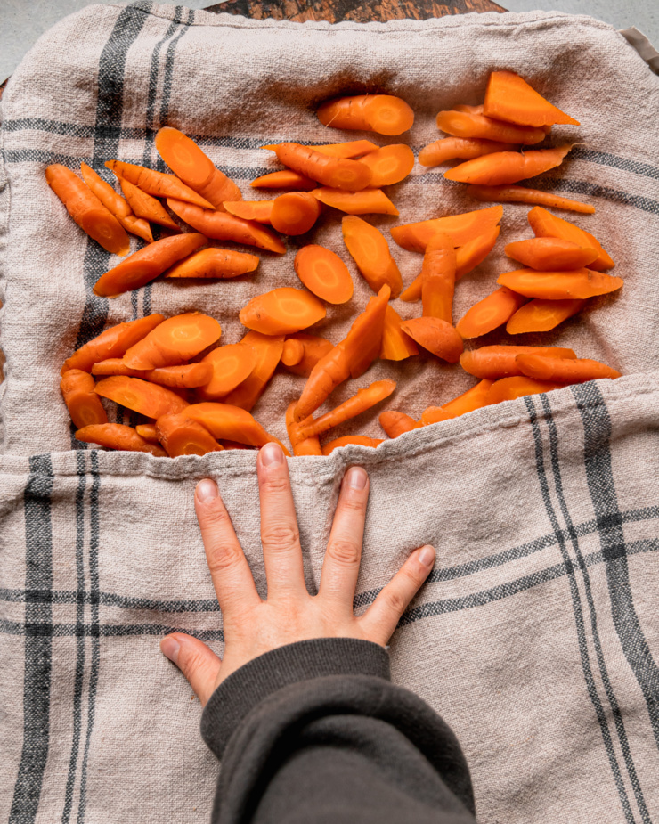 An overhead shot shows a hand using a kitchen towel to blot par-boiled carrots.