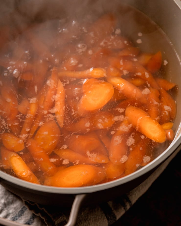 An overhead shot shows chopped carrots in a pot of boiling water. Steam is coming off the pot.