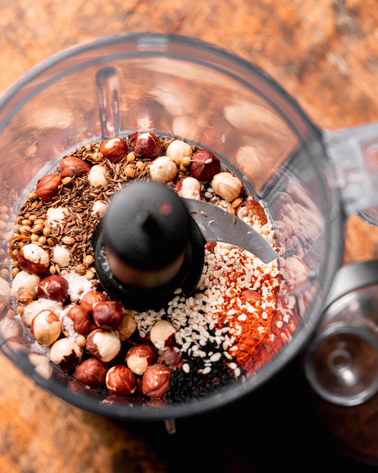 An overhead shot shows hazelnuts, spices, ground chilies, and sesame seeds in the bowl of a mini food processor.