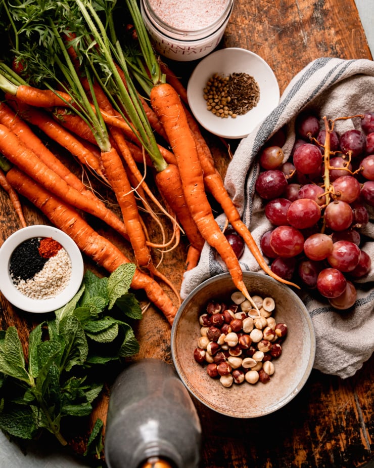 An overhead shot shows ingredients used in a roasted carrot side dish on a worn wood board: salt, carrots, red grapes, cumin seed, coriander seed, hazelnuts, sesame seeds, nigella seeds, ground chilies, mint, and olive oil.