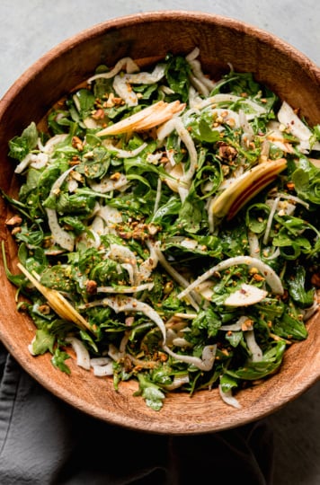 An overhead shot shows a shaved arugula fennel salad with apples and finely chopped pistachios. The salad is in a wooden bowl with a linen napkin nearby.