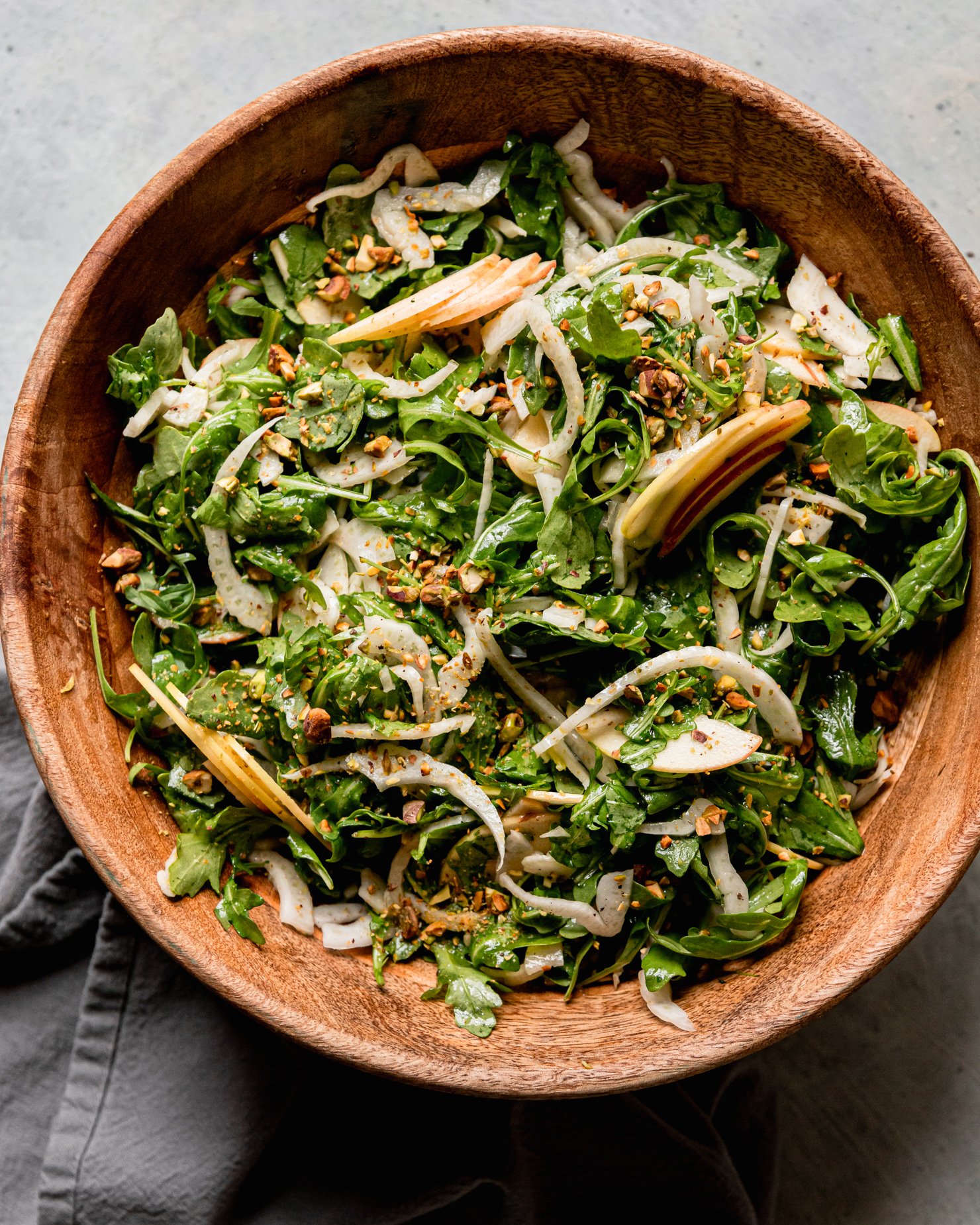 An overhead shot shows a shaved arugula fennel salad with apples and finely chopped pistachios. The salad is in a wooden bowl with a linen napkin nearby.