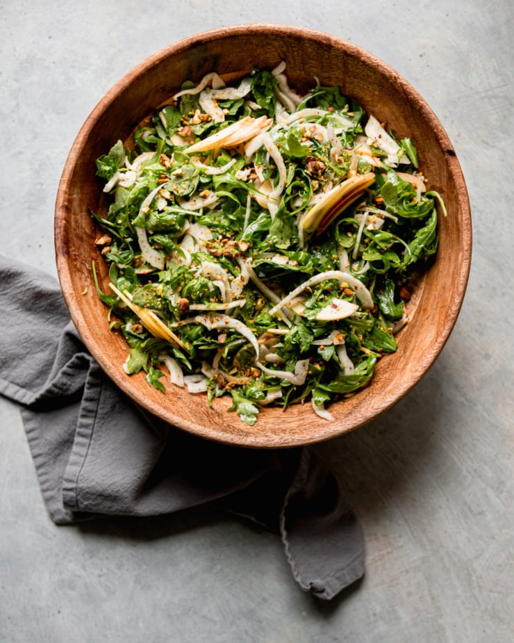 An overhead shot shows a shaved arugula fennel salad with apples and finely chopped pistachios. The salad is in a wooden bowl with a linen napkin nearby.