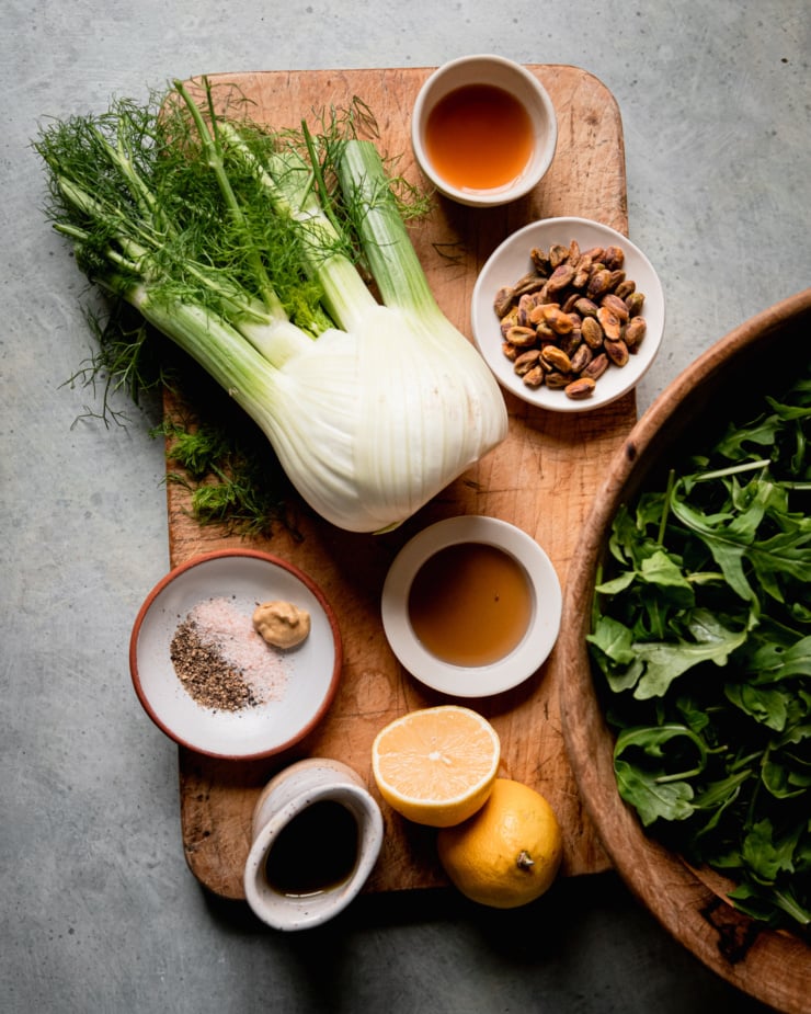 An overhead shot shows ingredients for a shaved arugula fennel salad: apple cider vinegar, pistachios, arugula, maple syrup, lemon, olive oil, salt, pepper, Dijon mustard, and fresh fennel.