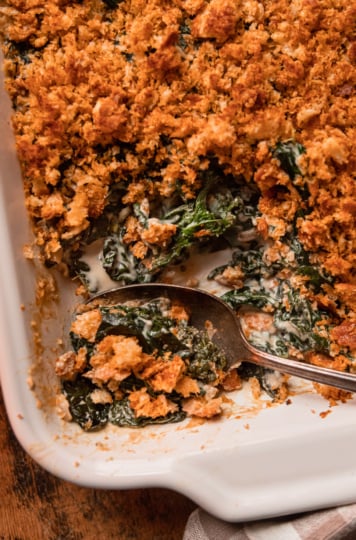 An up close, overhead shot shows a baked dish of vegan creamed kale with toasted bread crumb topping. A few scoops have been taken out of the dish and spoon sticks out at the bottom.