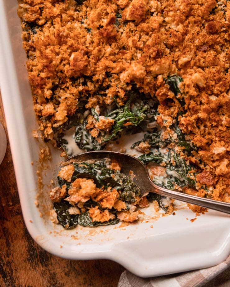 An up close, overhead shot shows a baked dish of vegan creamed kale with toasted bread crumb topping. A few scoops have been taken out of the dish and spoon sticks out at the bottom.