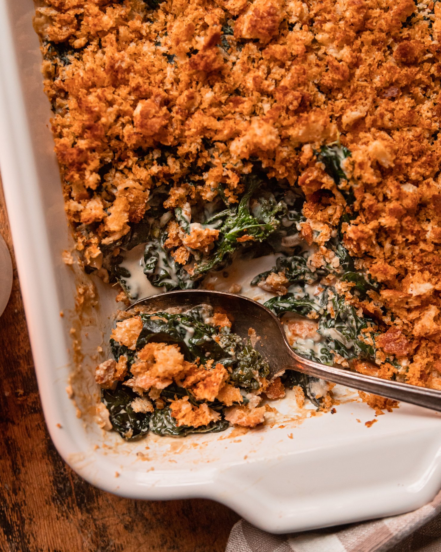An up close, overhead shot shows a baked dish of vegan creamed kale with toasted bread crumb topping. A few scoops have been taken out of the dish and spoon sticks out at the bottom.