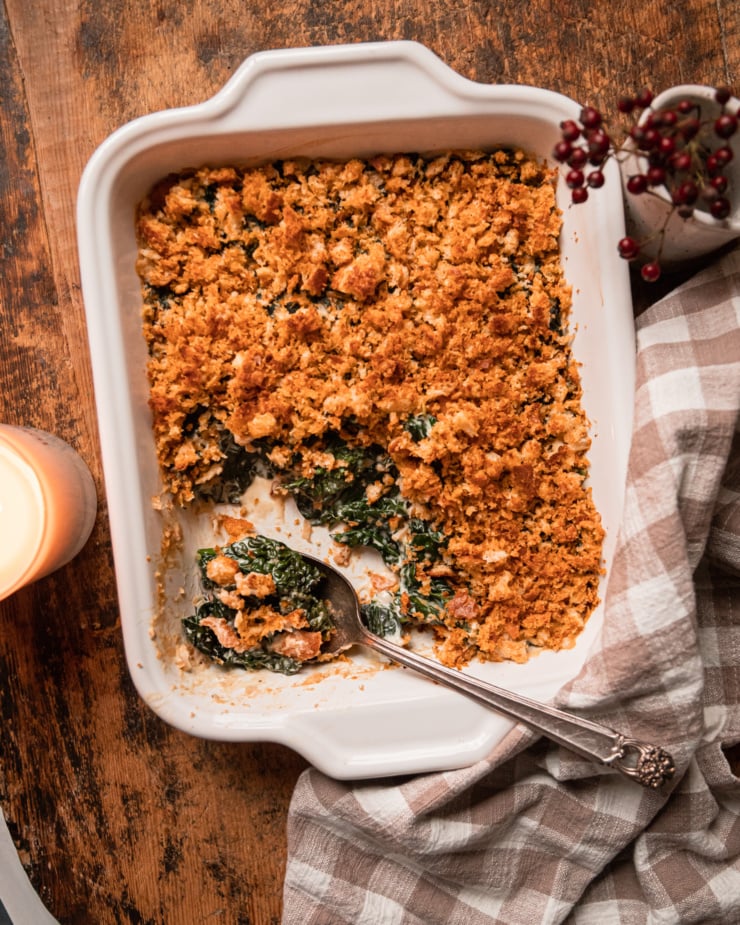 An overhead shot shows a baked dish of vegan creamed kale with toasted bread crumb topping. A few scoops have been taken out of the dish and spoon sticks out at the bottom.