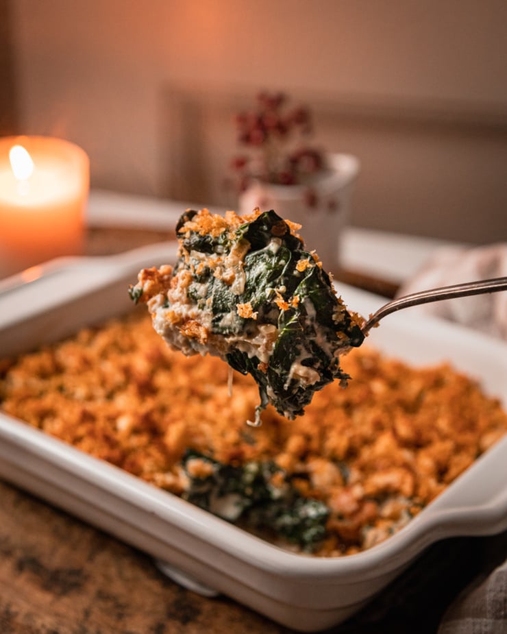 An up close shot shows a big spoonful of vegan creamed kale with some toasted bread crumbs on top. The whole baking dish of kale is in the background, as well as a lit candle.