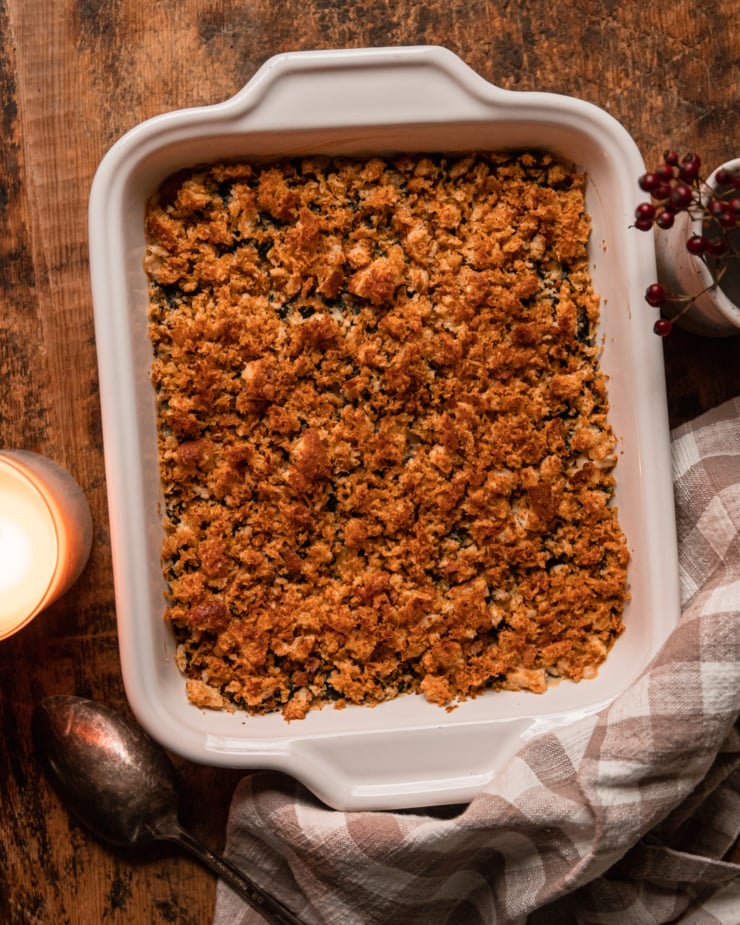 An overhead shot shows a baked dish of vegan creamed kale with bread crumb topping on a worn wood background.