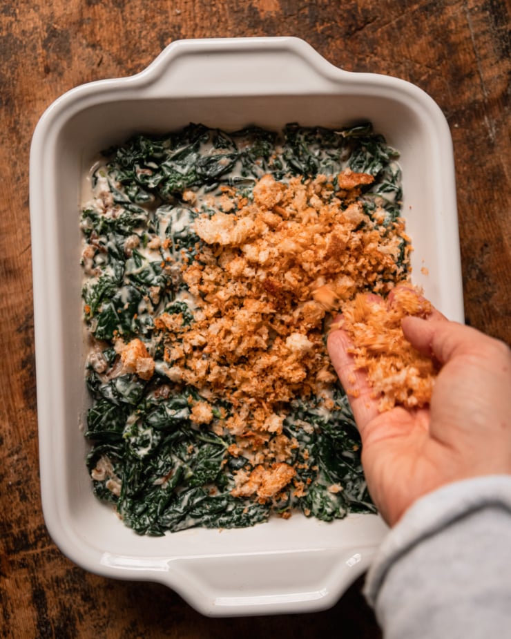 An overhead shot shows a baking dish filled with vegan creamed kale. A hand is placing bread crumbs on top of the creamed kale.