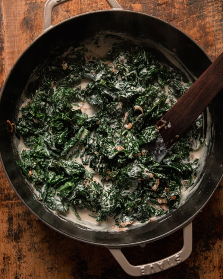 An overhead shot shows a pan filled with creamy cooked kale with a savoury cashew cream. A wooden utensil is sticking out of the pan.