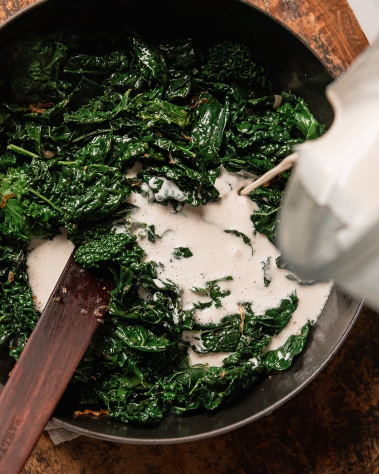 An overhead shot shows a pan filled with wilted kale. A cashew "cream" is being poured into the pan.