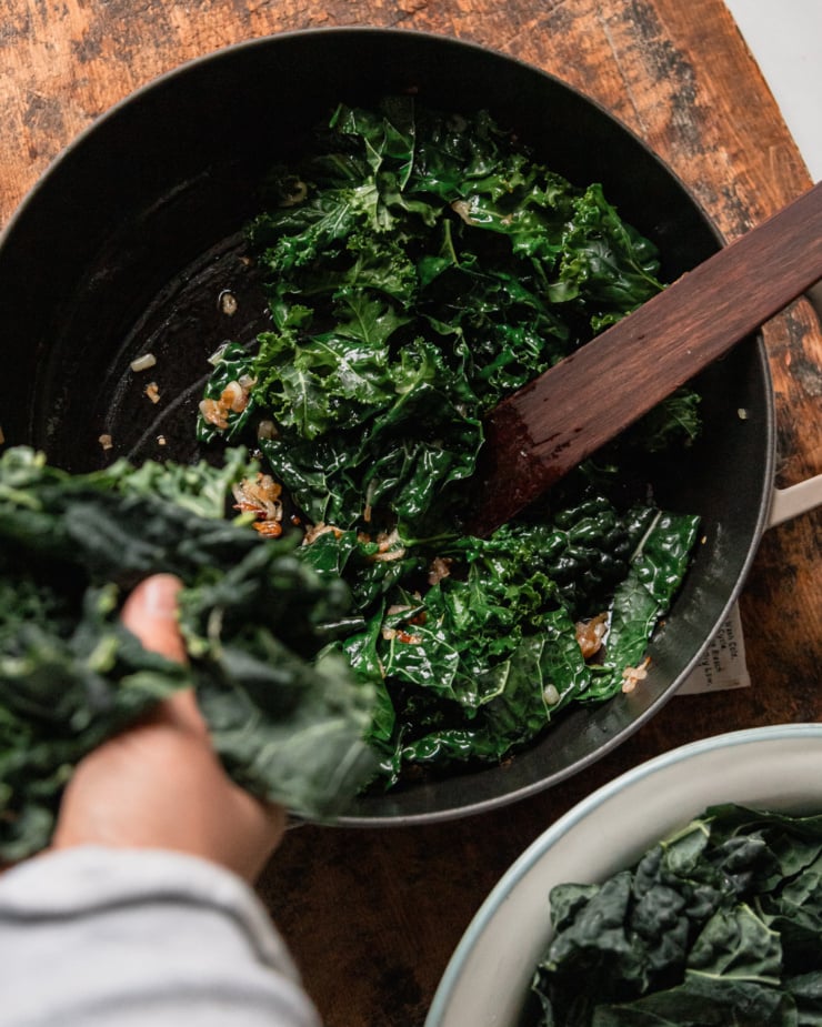 An overhead shot shows a hand adding more chopped kale to a pan with wilted kale and caramelized shallots.