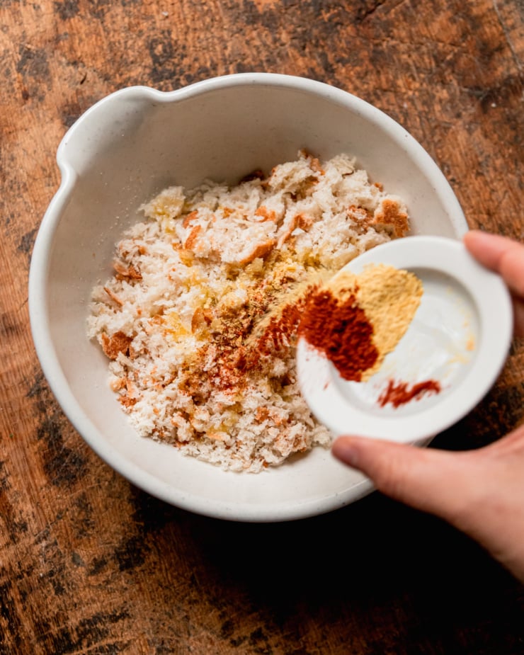 An overhead shot shows a hand emptying a bowl of spices into a bowl of fresh bread crumbs.