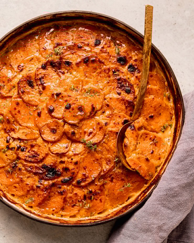 An overhead shot shows a vegan sweet potato gratin in a ceramic baking dish. The top is broiled and browned in spots. A brass serving spoon is sticking out of the gratin.