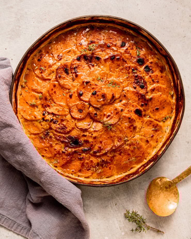 An overhead shot shows a vegan sweet potato gratin in a ceramic baking dish. The top is broiled and browned in spots. A brass serving spoon is nearby.