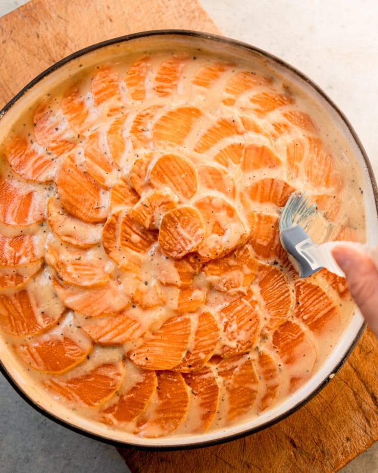 An overhead shot shows a hand using a silicone pastry brush to distribute a cashew "cream" mixture over baked sweet potato slices in a dish.