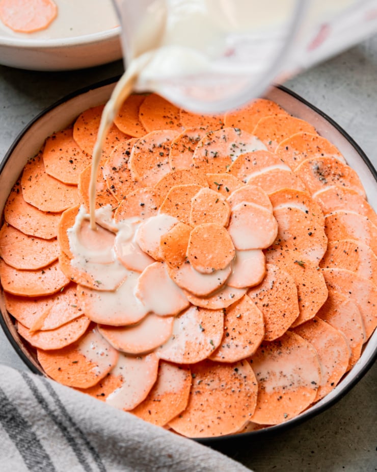 A slight 3/4 angle shot shows cashew "cream" being poured on top of layered sweet potato slices in a baking dish, arranged in a pretty spiral pattern.