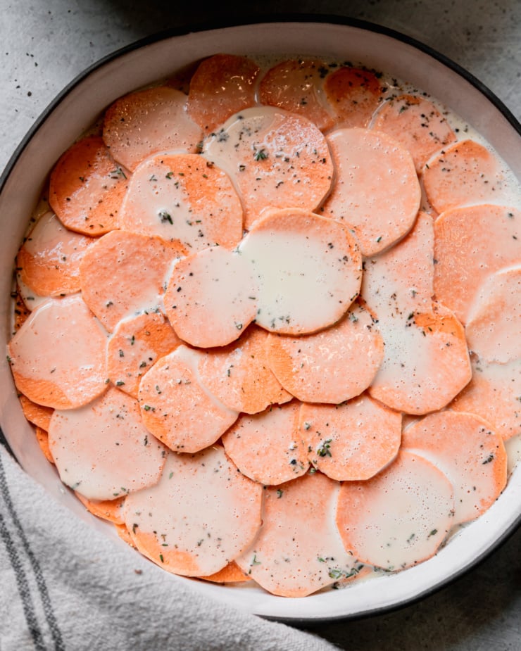 An overhead shot shows a ceramic baking dish filled with round sweet potato slices. There is a layer of cashew "cream" on top as well as salt, pepper, and minced thyme.