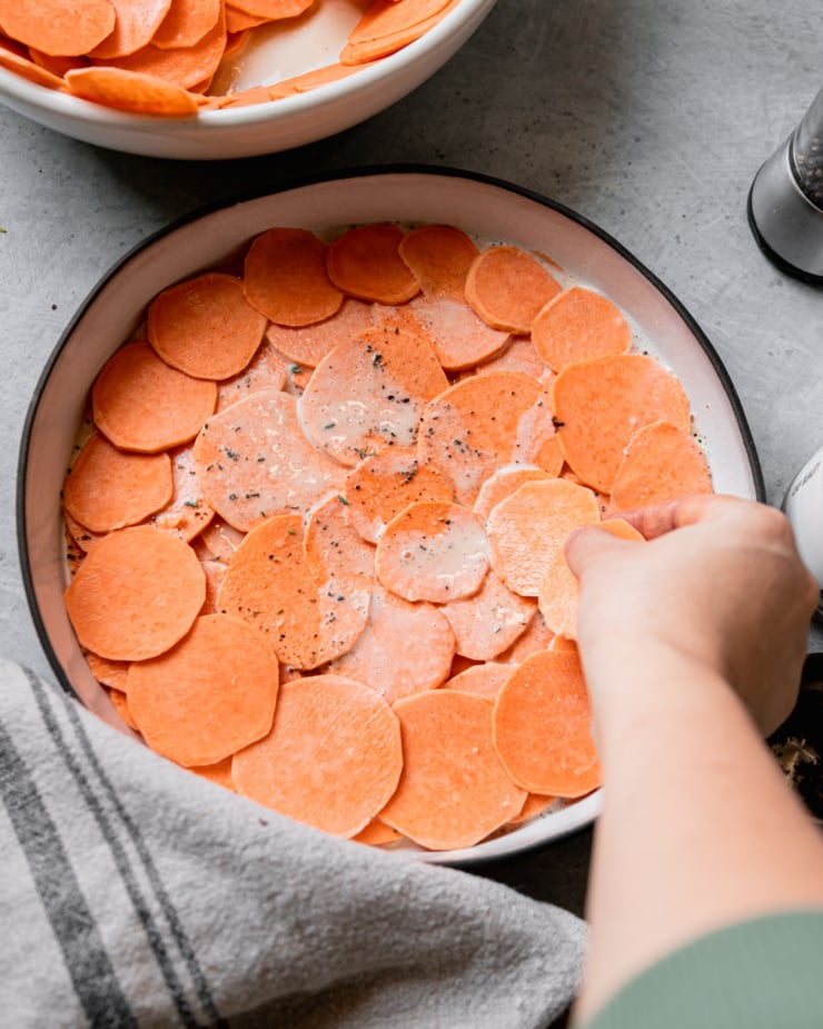 An overhead shot shows a hand layering sweet potato slices into a baking dish.