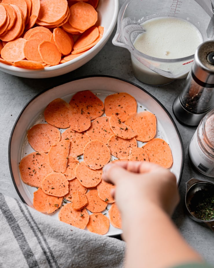An overhead shot shows sweet potato slices in a ceramic baking dish. A hand is sprinkling minced thyme leaves on top.