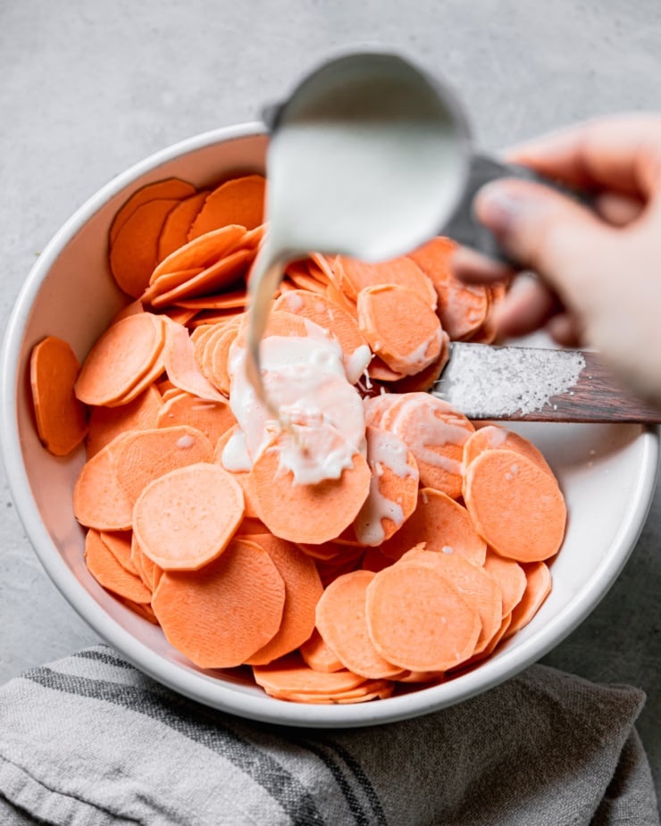 An overhead shot shows a hand using a measuring cup to pour cashew "cream" on top of a bowl of sliced sweet potatoes.