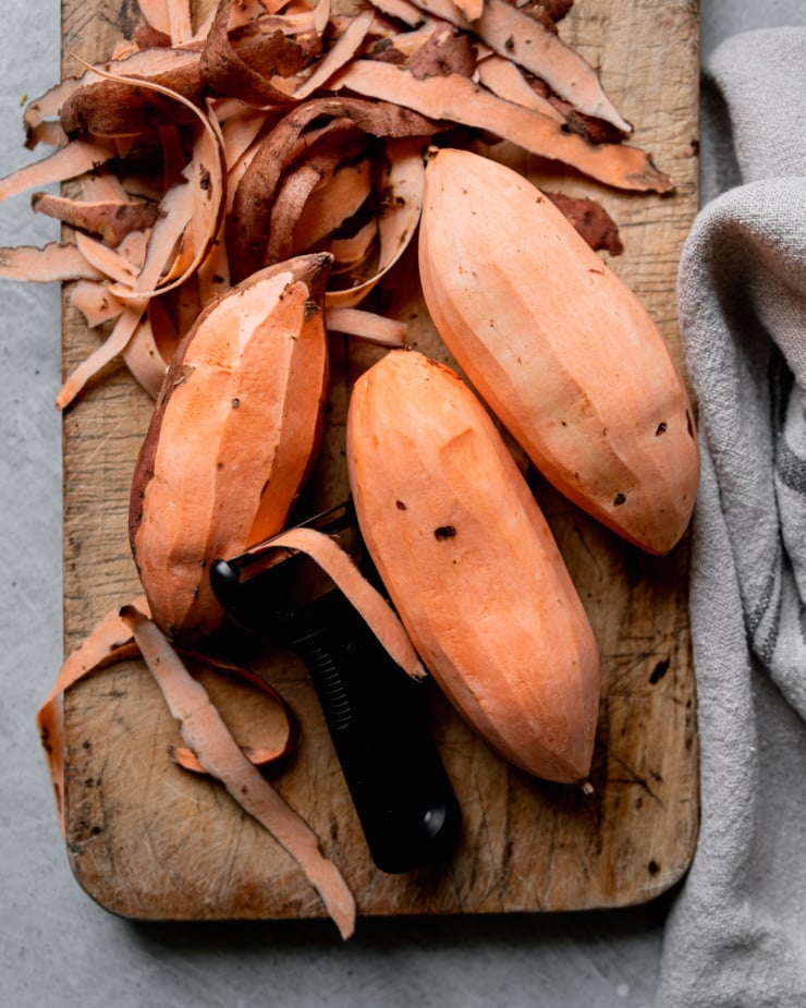 An overhead shot shows peeled sweet potatoes on a worn wood cutting board.
