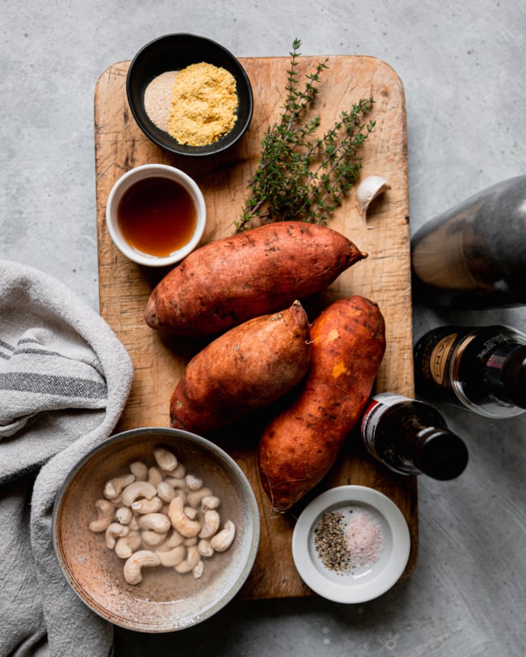 An overhead shot shows ingredients for a vegan sweet potato gratin: sweet potatoes, maple syrup, nutritional yeast, onion powder, raw cashews, liquid smoke, Tamari, olive oil, salt, pepper, thyme, and garlic.