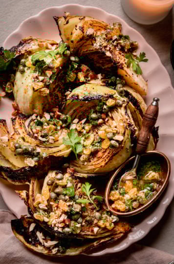 An up close, overhead shot shows a platter with well-browned and caramelized roasted cabbage wedges. The wedges are topped with a caper and golden raisin relish mixture, chopped parsley, and almonds.