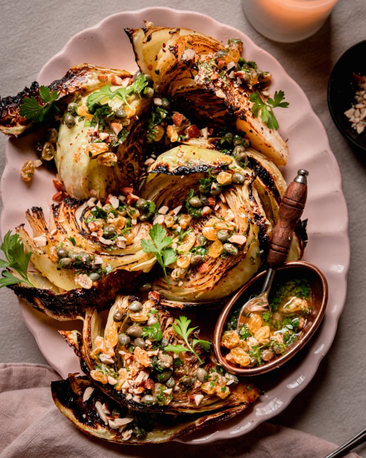 An up close, overhead shot shows a platter with well-browned and caramelized roasted cabbage wedges. The wedges are topped with a caper and golden raisin relish mixture, chopped parsley, and almonds.