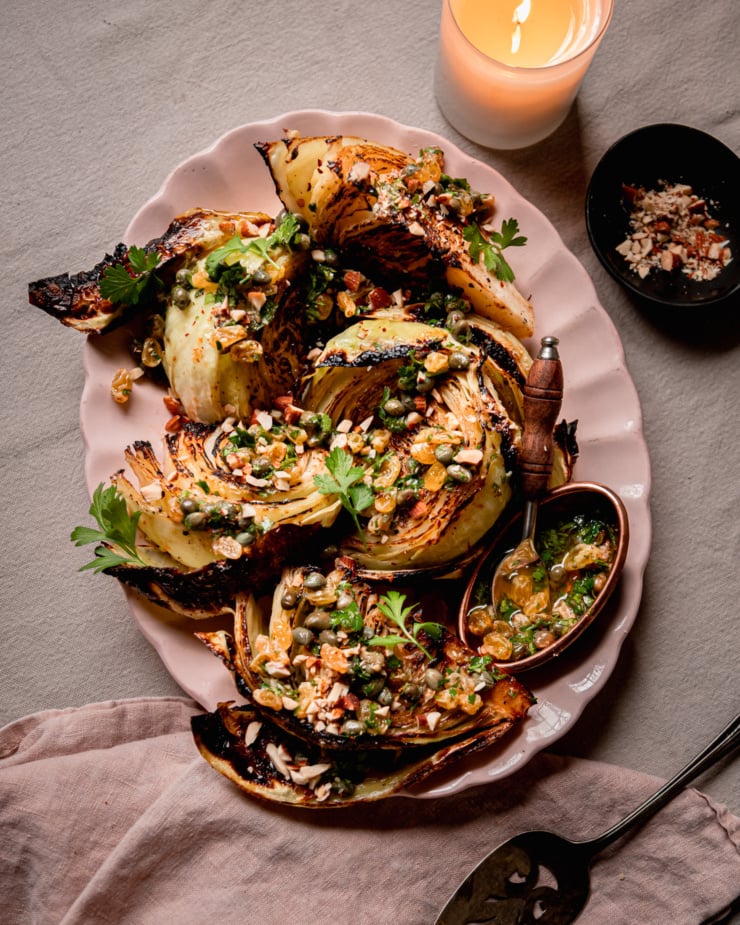 An overhead shot shows a platter with well-browned and caramelized roasted cabbage wedges. The wedges are topped with a caper and golden raisin relish mixture, chopped parsley, and almonds. A small bowl of chopped almonds and a lit candle is nearby.