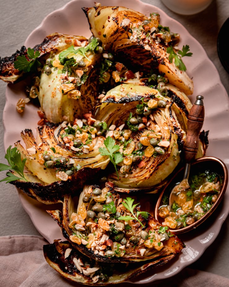 An up close, overhead shot shows a platter with well-browned and caramelized roasted cabbage wedges. The wedges are topped with a caper and golden raisin relish mixture, chopped parsley, and almonds.