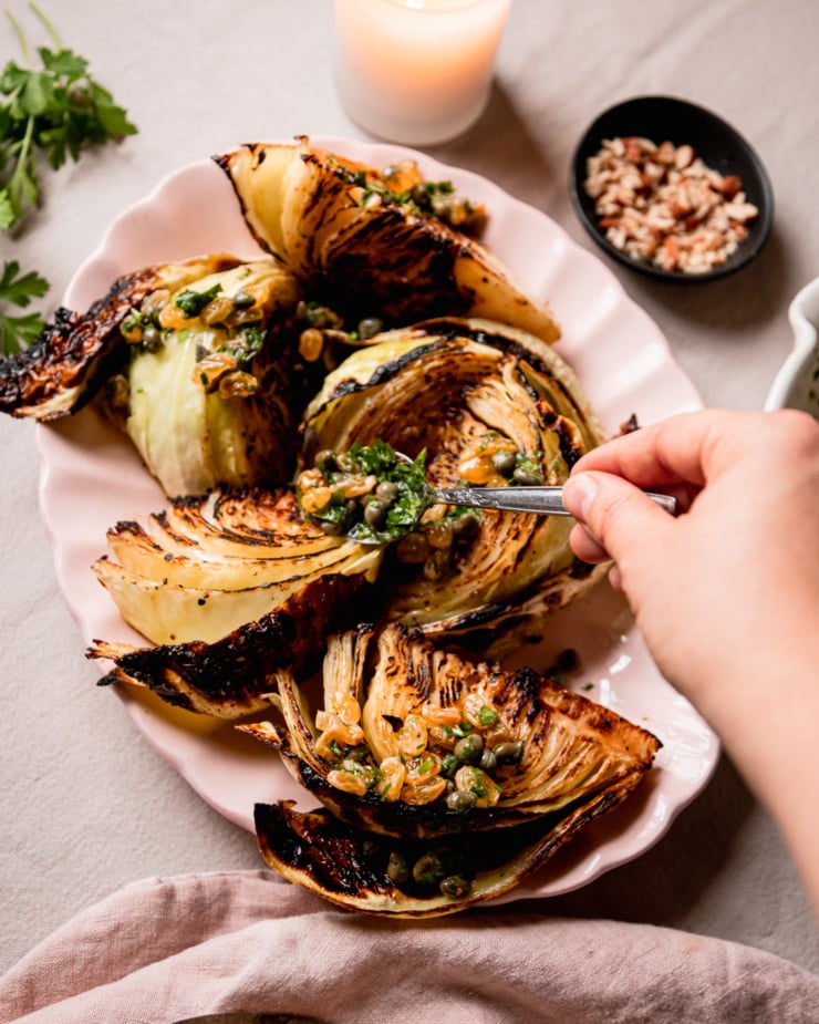 An overhead shot shows a hand using a spoon to distribute caper raisin relish over roasted cabbage wedges.
