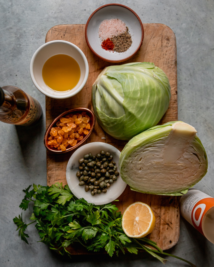 An overhead shot shows ingredients for a vegetable side dish: cabbage, capers, golden raisins, lemon, sherry vinegar, parsley, vegan Worcestershire sauce, olive oil, salt, pepper, and ground chilies.