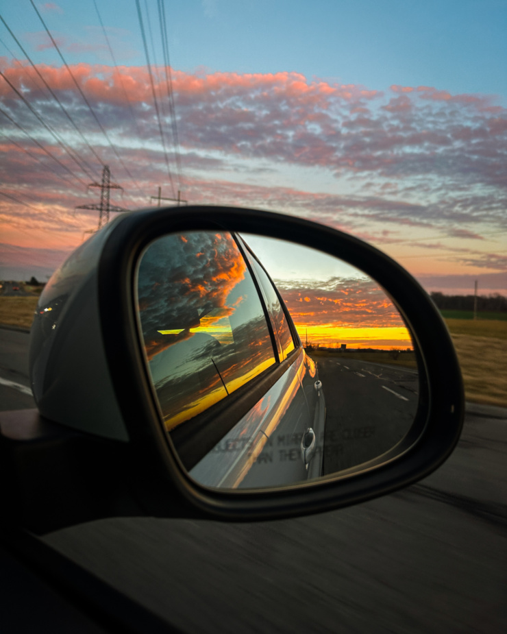 An up close shot shows a sunset in a car's rearview side mirror.