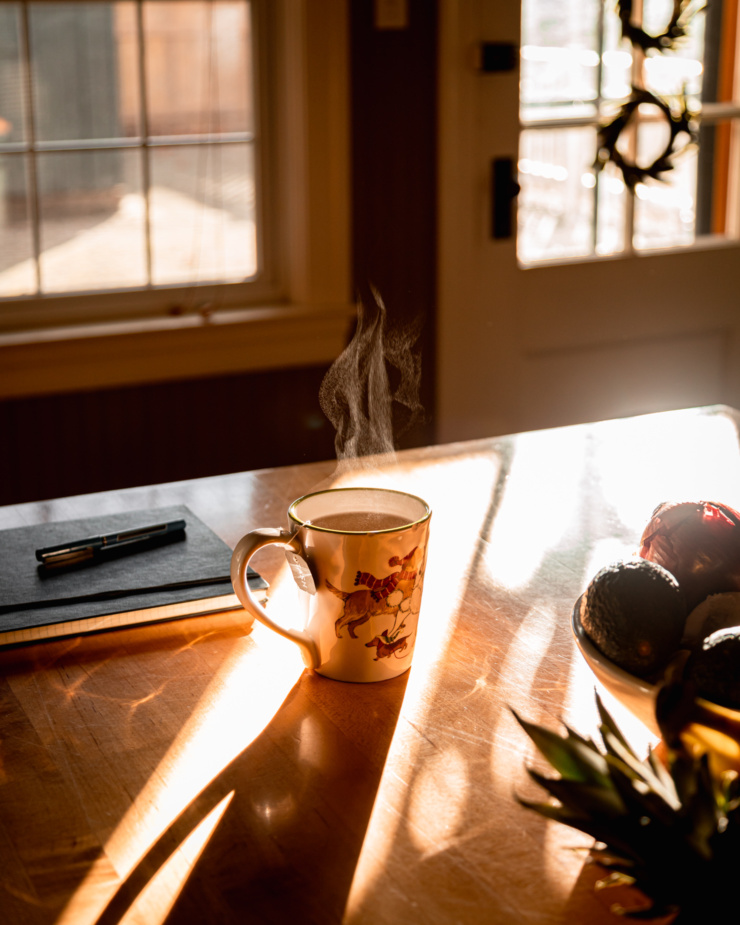 A head-on shot shows a steaming mug of tea on a kitchen counter in direct sunlight. A notebook and pen is nearby.
