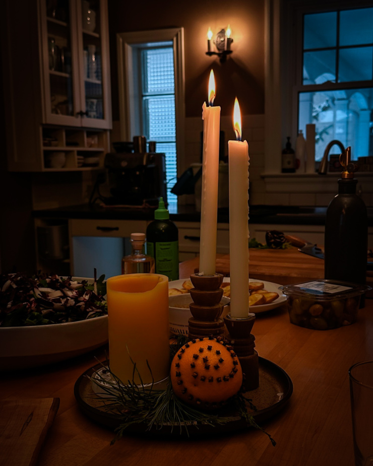 A head-on shot shows two lit candles on a plate with a pomander in a dimly lit kitchen.