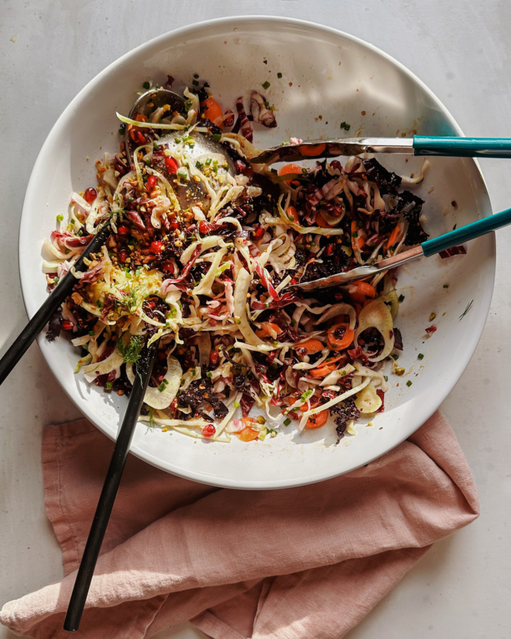 An overhead shot shows a shaved fennel and cabbage salad with pomegranate seeds. Two sets of tongs are in the bowl and half of the salad has been served.