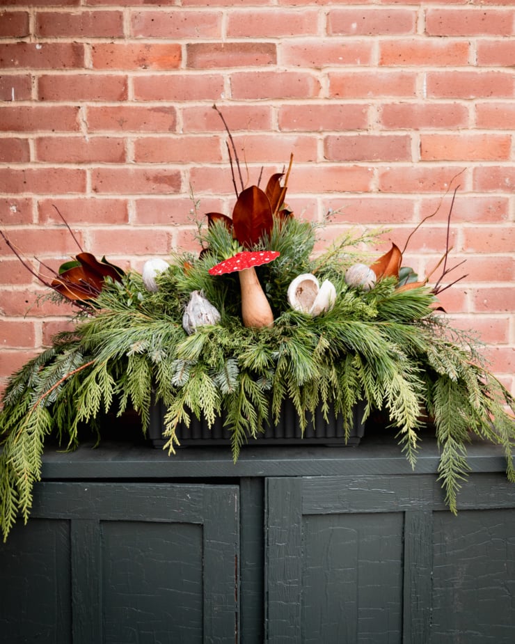 A head-on shot shows a holiday evergreen planter against a brick background outside. The planter features green and little pod/shell/mushroom ornaments.