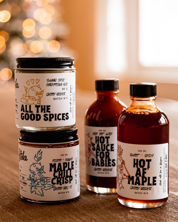 A head-on shot shows jars and bottles of hot sauce, spice blends, chili crisp, and spicy maple syrup. Twinkling lights are seen in the background.