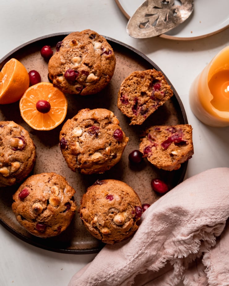 An overhead shot shows vegan cranberry clementine muffins with white chocolate on a speckled plate. A halved clementine and fresh cranberries are also on the plate. A lit candle is nearby.