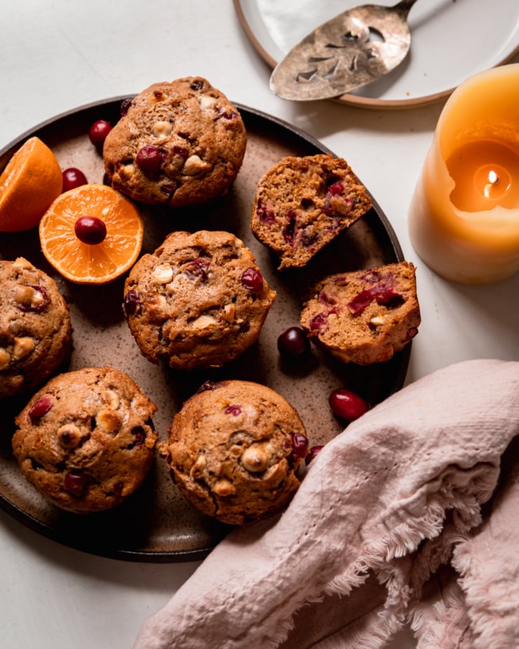 An overhead shot shows vegan cranberry clementine muffins with white chocolate on a speckled plate. A halved clementine and fresh cranberries are also on the plate. A lit candle is nearby.