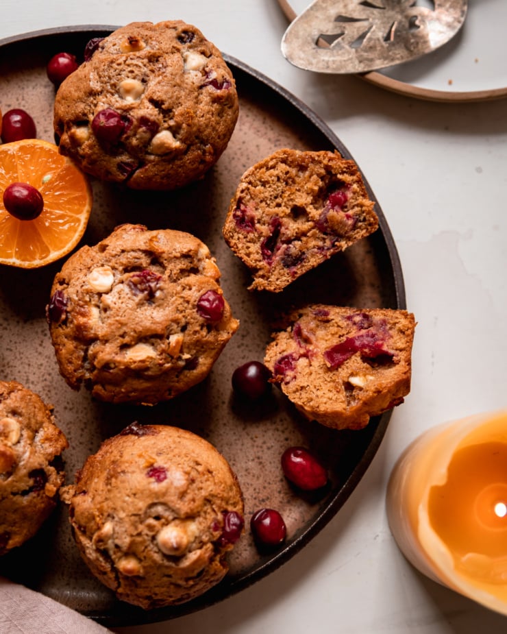 An overhead shot shows vegan cranberry clementine muffins with white chocolate on a speckled plate. A lit candle is nearby.