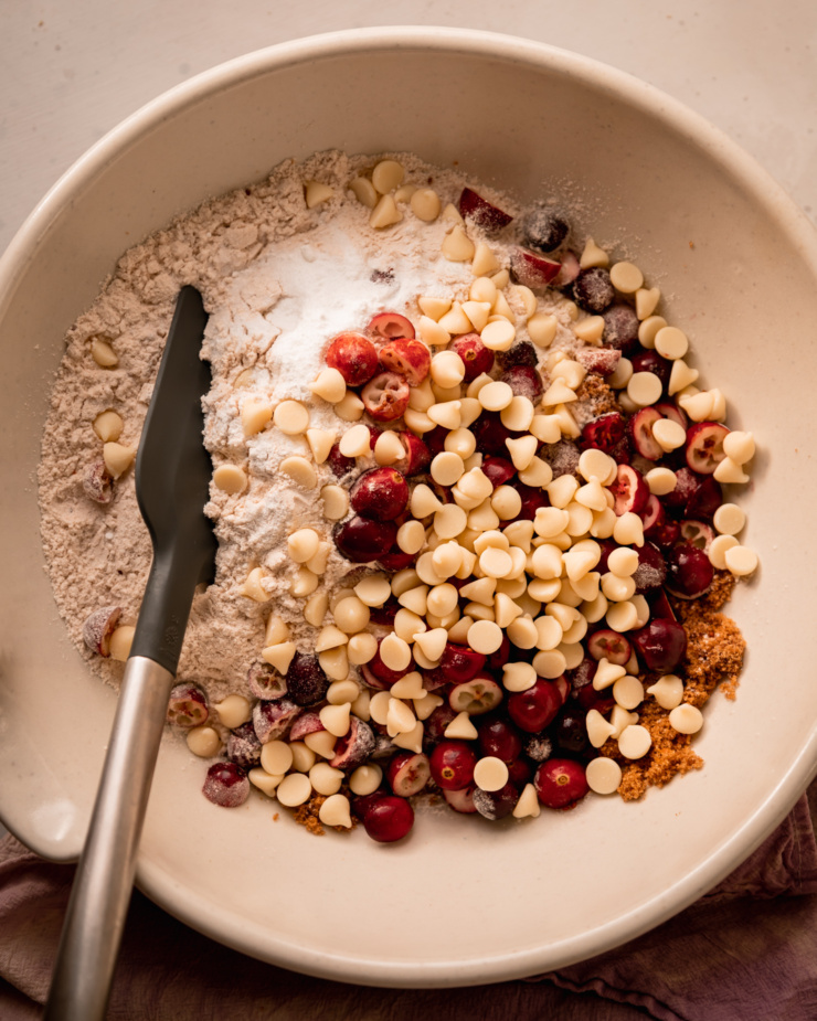 An overhead shot shows a flour mixture in a large bowl with chopped fresh cranberries and white chocolate chips.