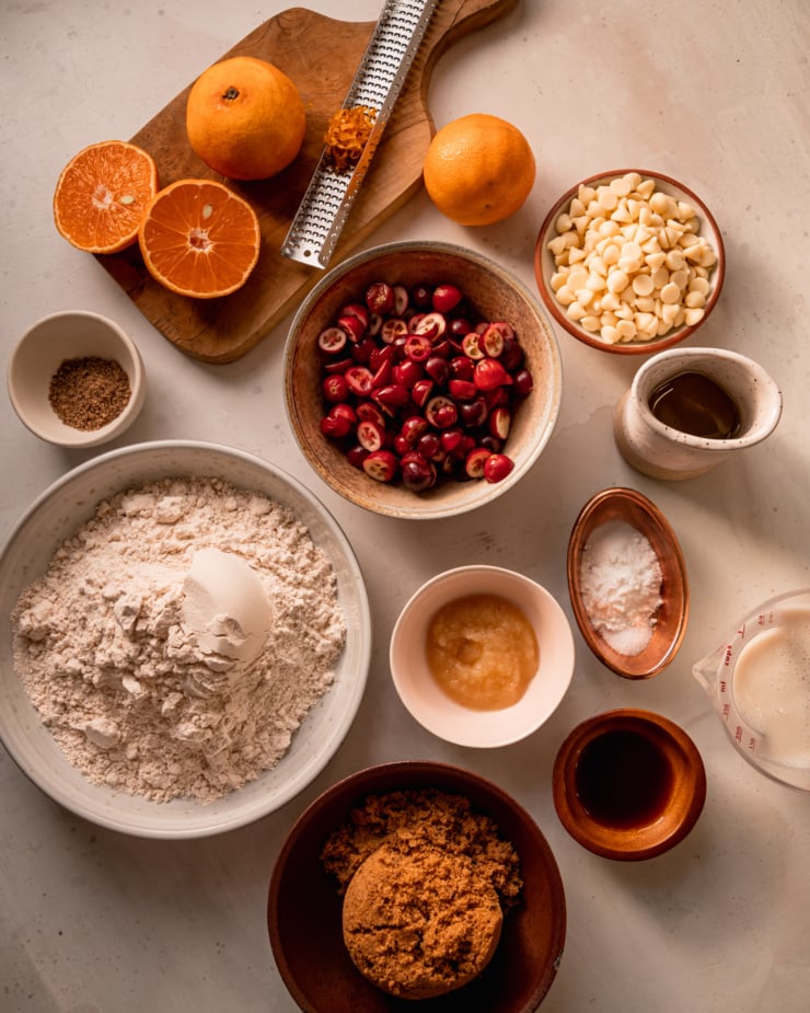 An overhead shot shows some ingredients for a batch of vegan muffins: cranberries, clementine zest and juice, brown sugar, light spelt flour, avocado oil, apple sauce, white chocolate chips, vanilla, and ground flax.