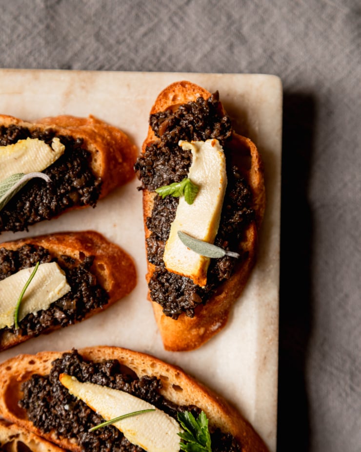 An up close, overhead shot focuses on a singular vegan mushroom duxelles crostini. The duxelles is like a dark brown paste and it is garnished with a small wedge of creamy vegan cheese and a couple herb leaves.